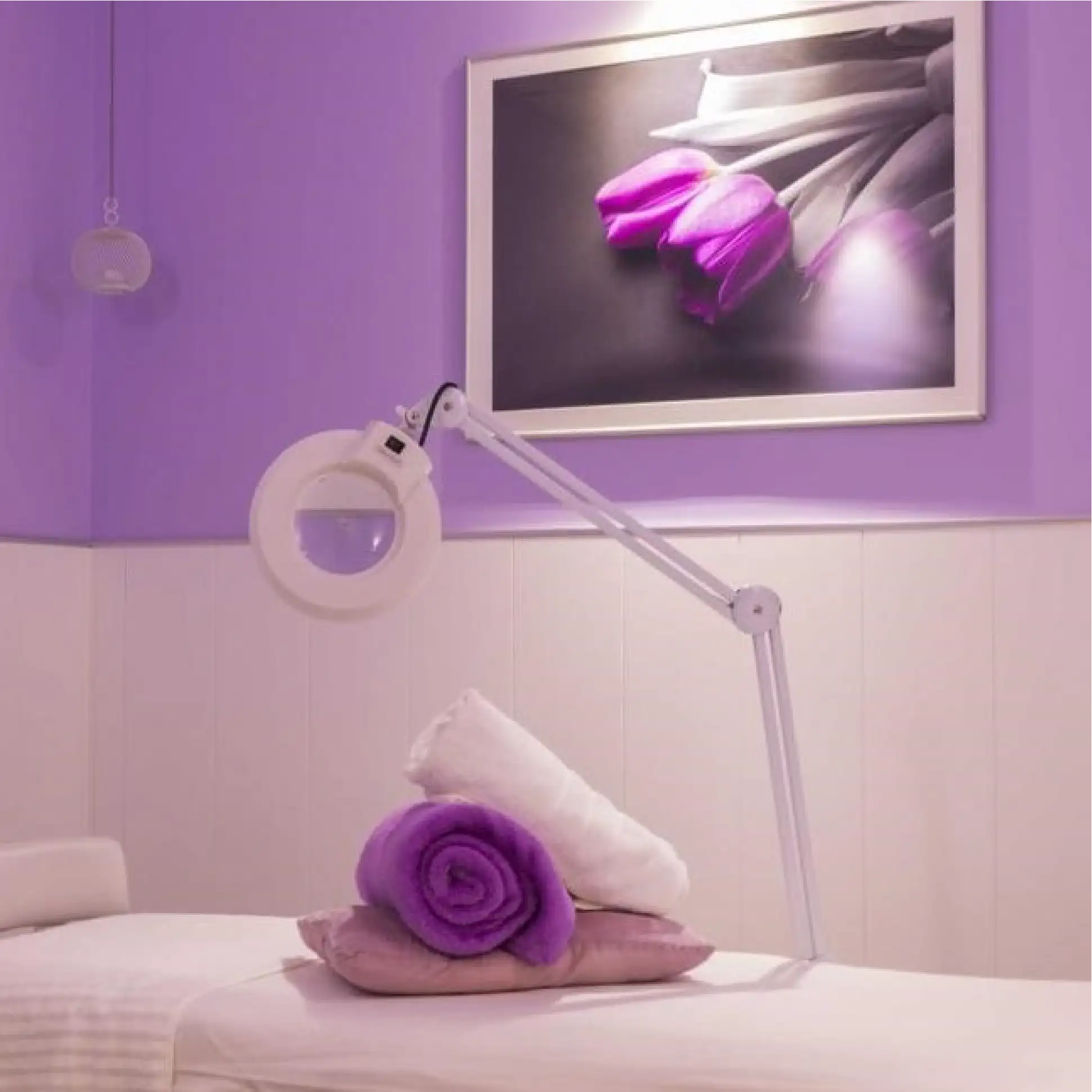 Interior de una sala de tratamiento con una camilla, toallas y una lámpara con lupa para tratamientos faciales.