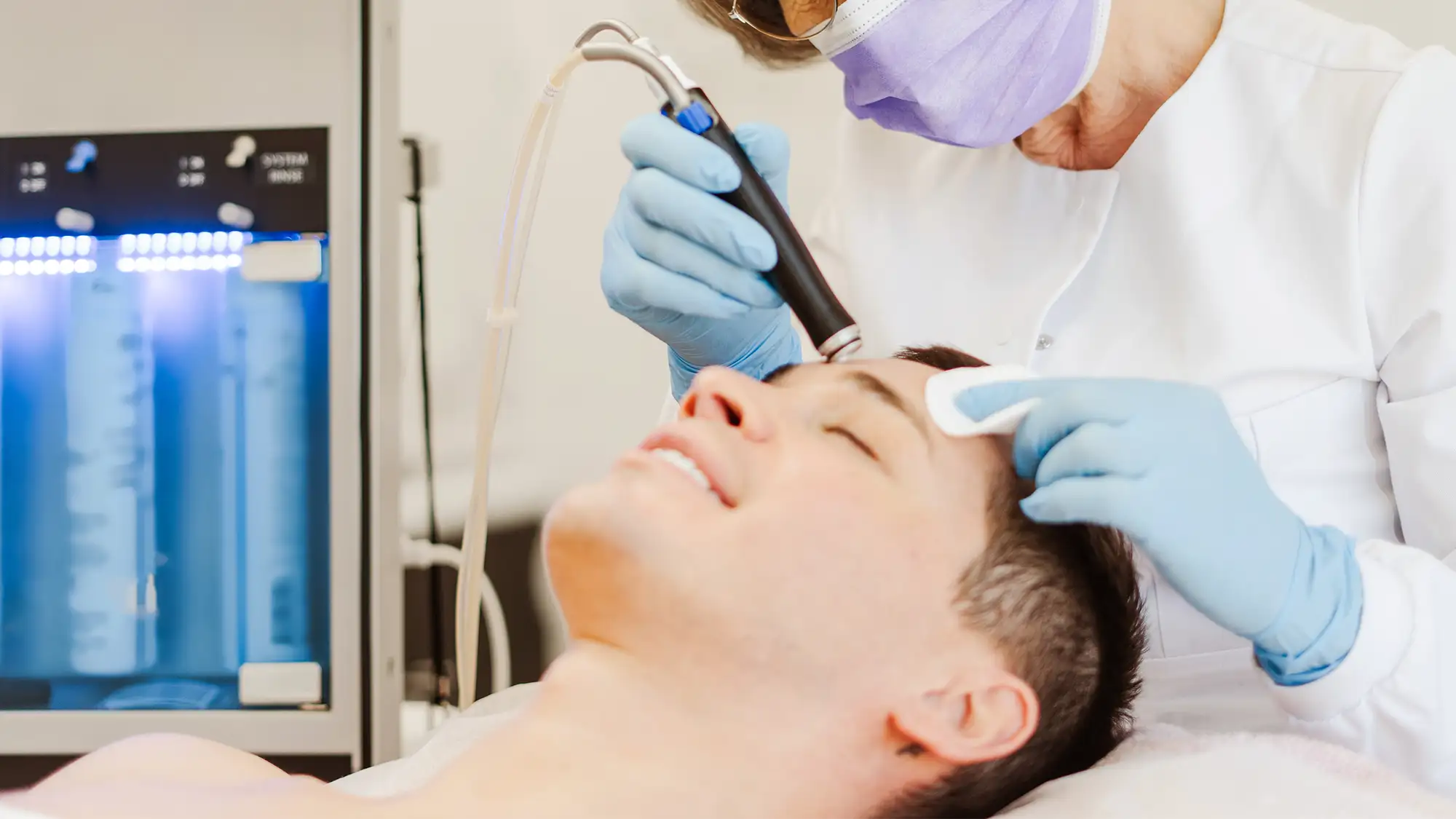 Paciente recibiendo tratamiento facial con dispositivo de hidrodermoabrasión en clínica estética, atendido por profesional con guantes y mascarilla.